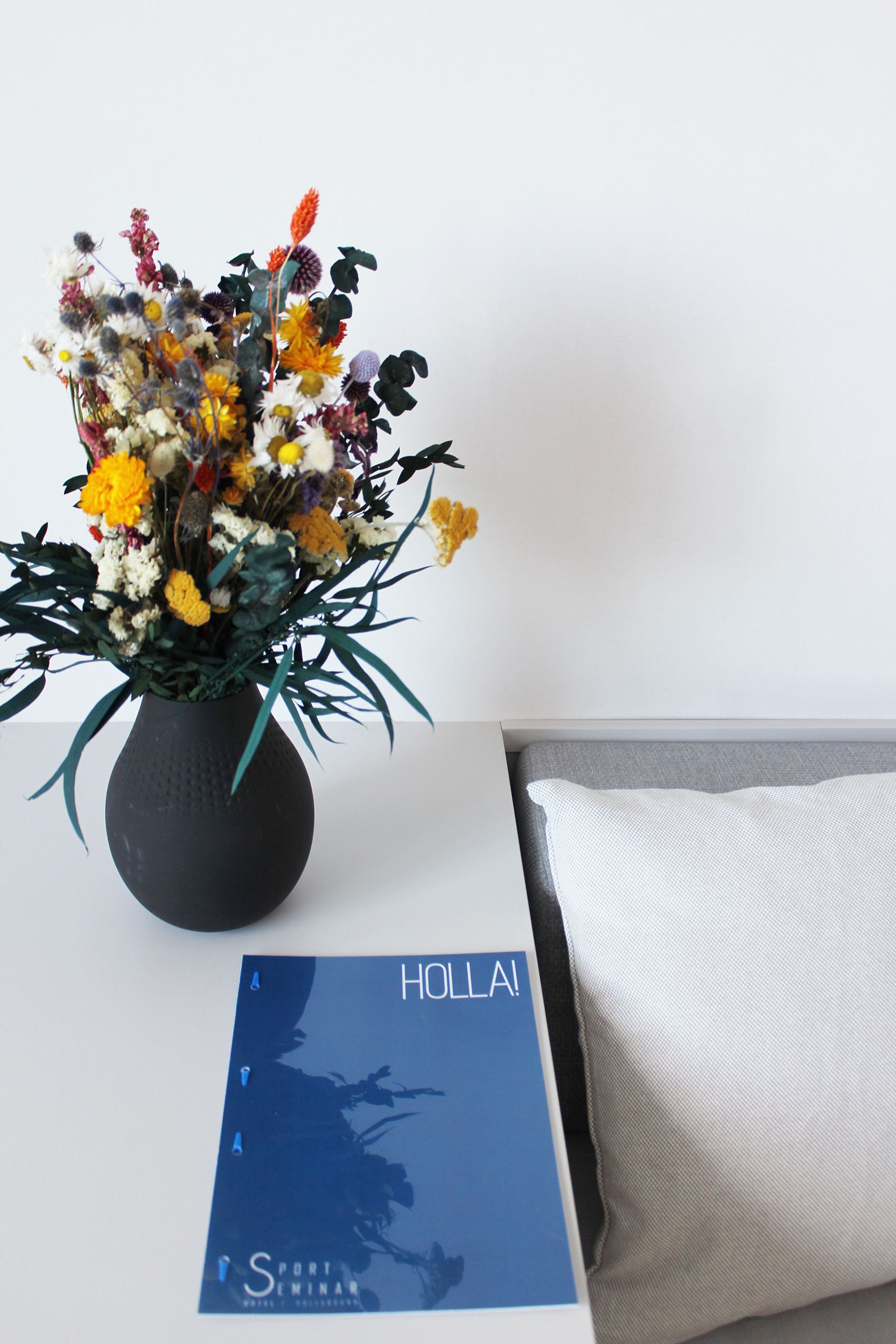 A table with a black vase full of colorful flowers, a blue magazine with the inscription 'HOLLA!' and a white cushion.