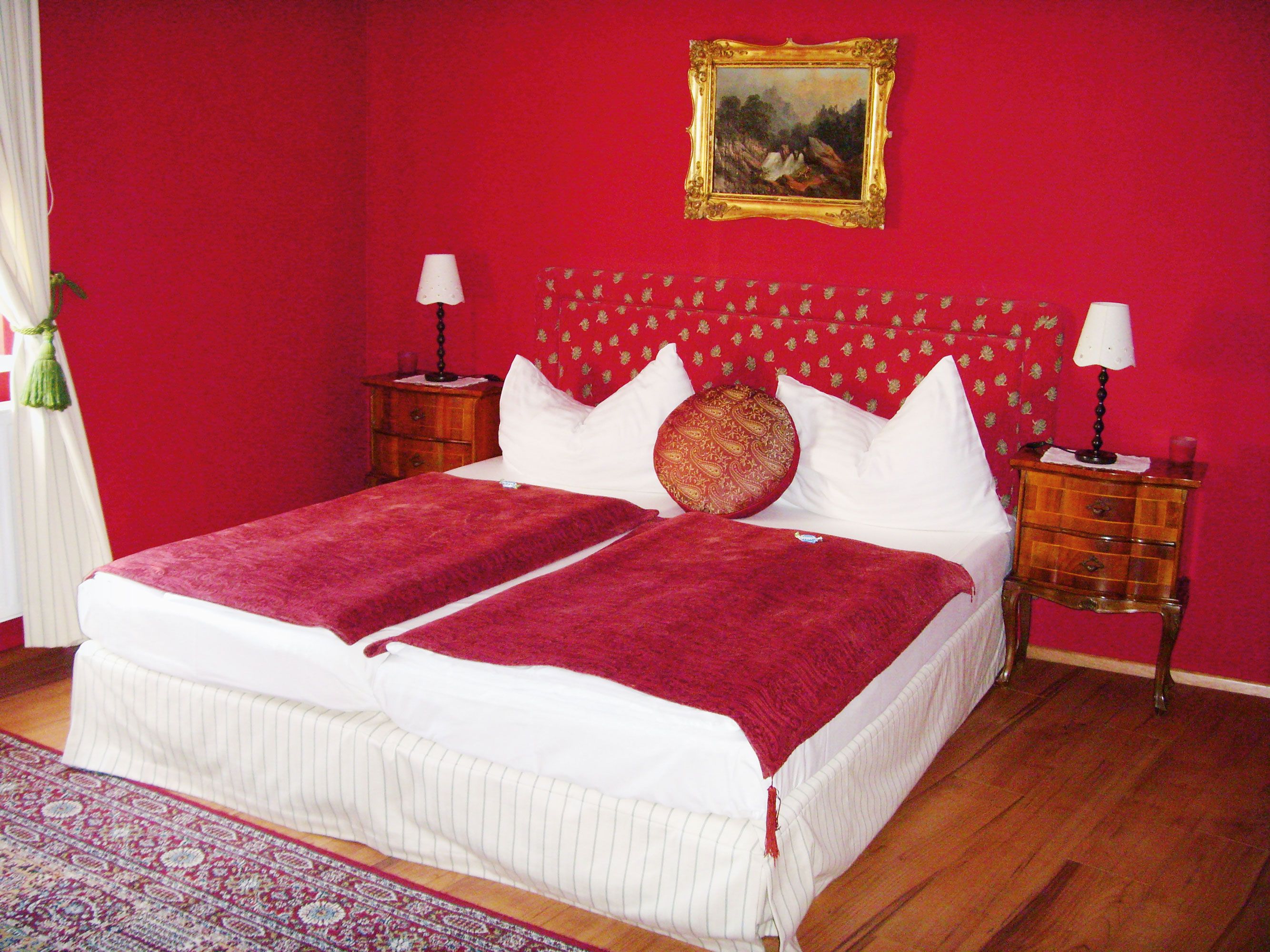 A bedroom with red walls, a double bed with red blankets and a painting on the wall.