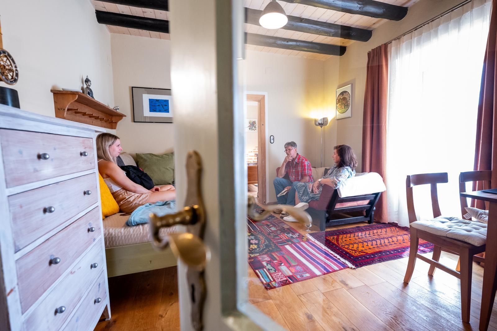 Three people sit in a cozy living room with a wooden floor and colorful carpet.