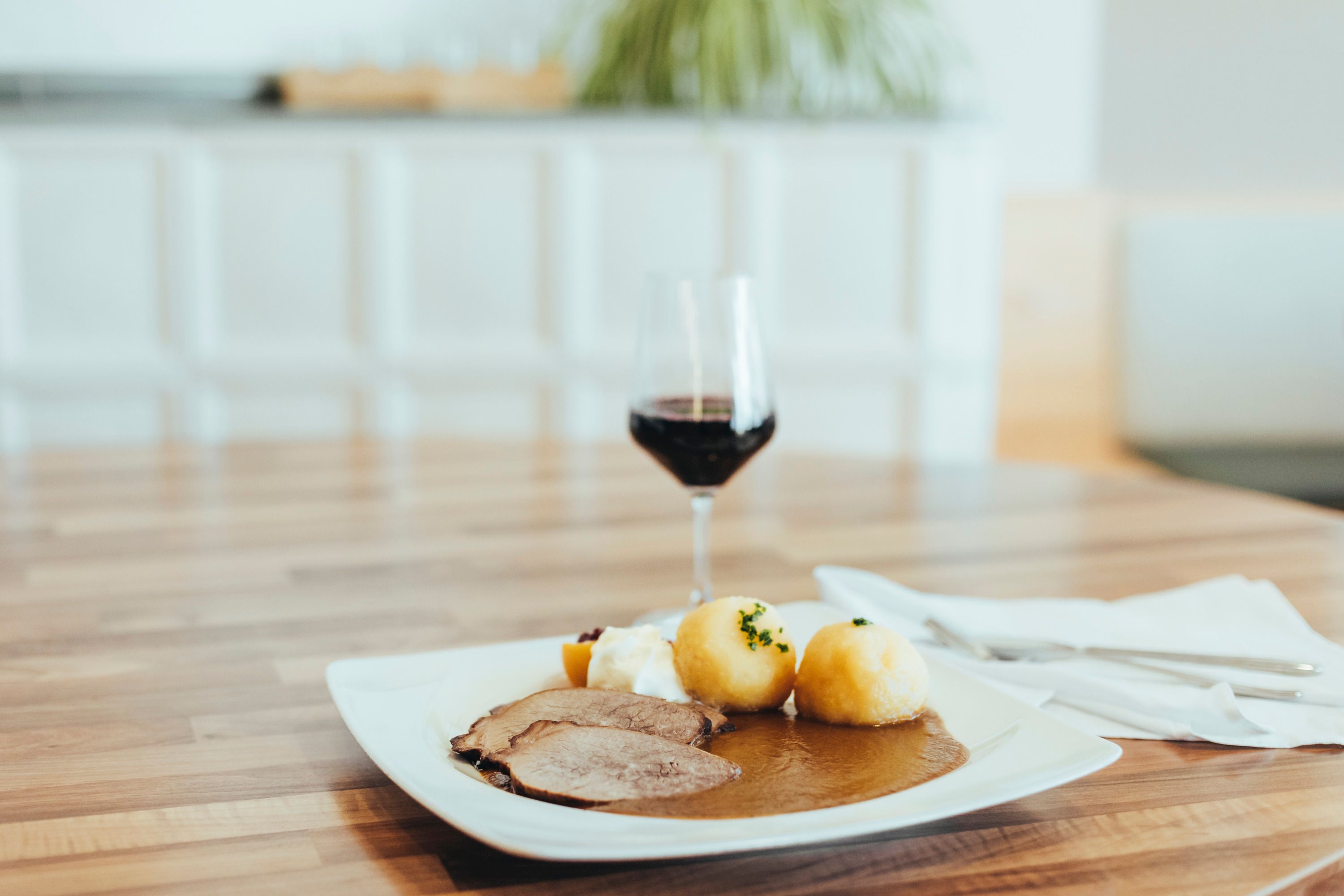 A plate of roast beef, dumplings and gravy on a wooden table, with a glass of red wine next to it.