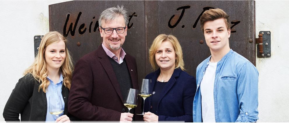 Four people stand in front of a wine cellar door holding glasses of white wine; friendly group shot with natural light and a familiar atmosphere.