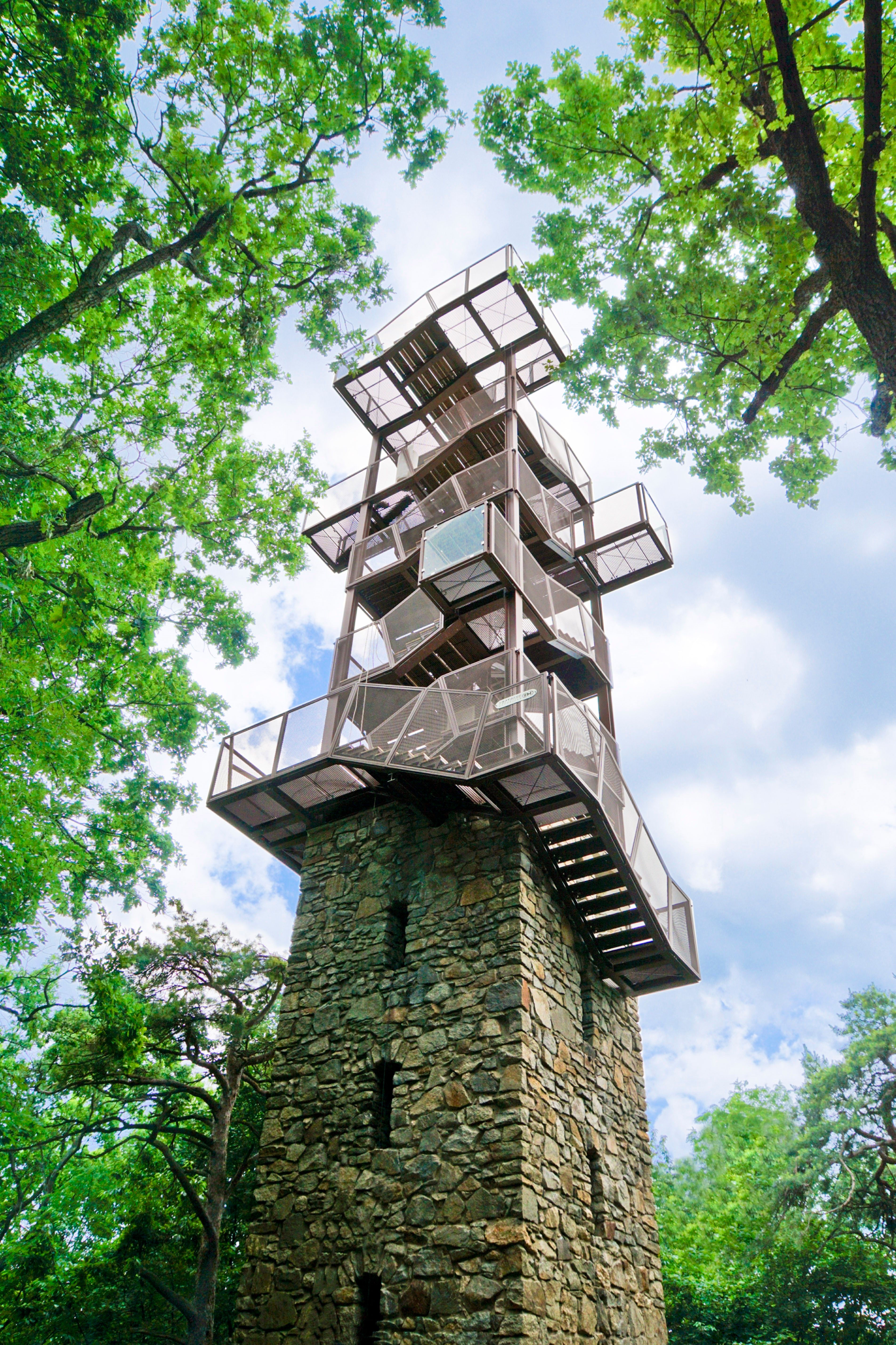 A modern observation tower made of metal and stone, surrounded by green trees, rises into the blue sky.