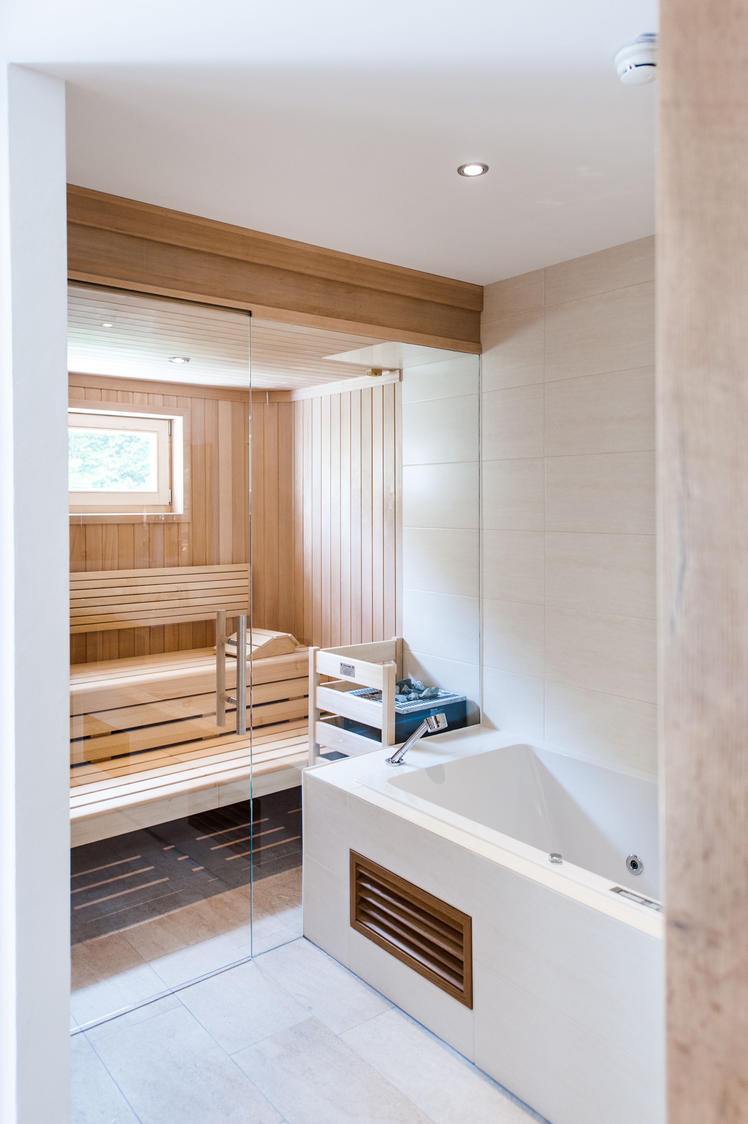 Interior view of a modern sauna with wooden paneling and glass wall, next to a white bathtub.
