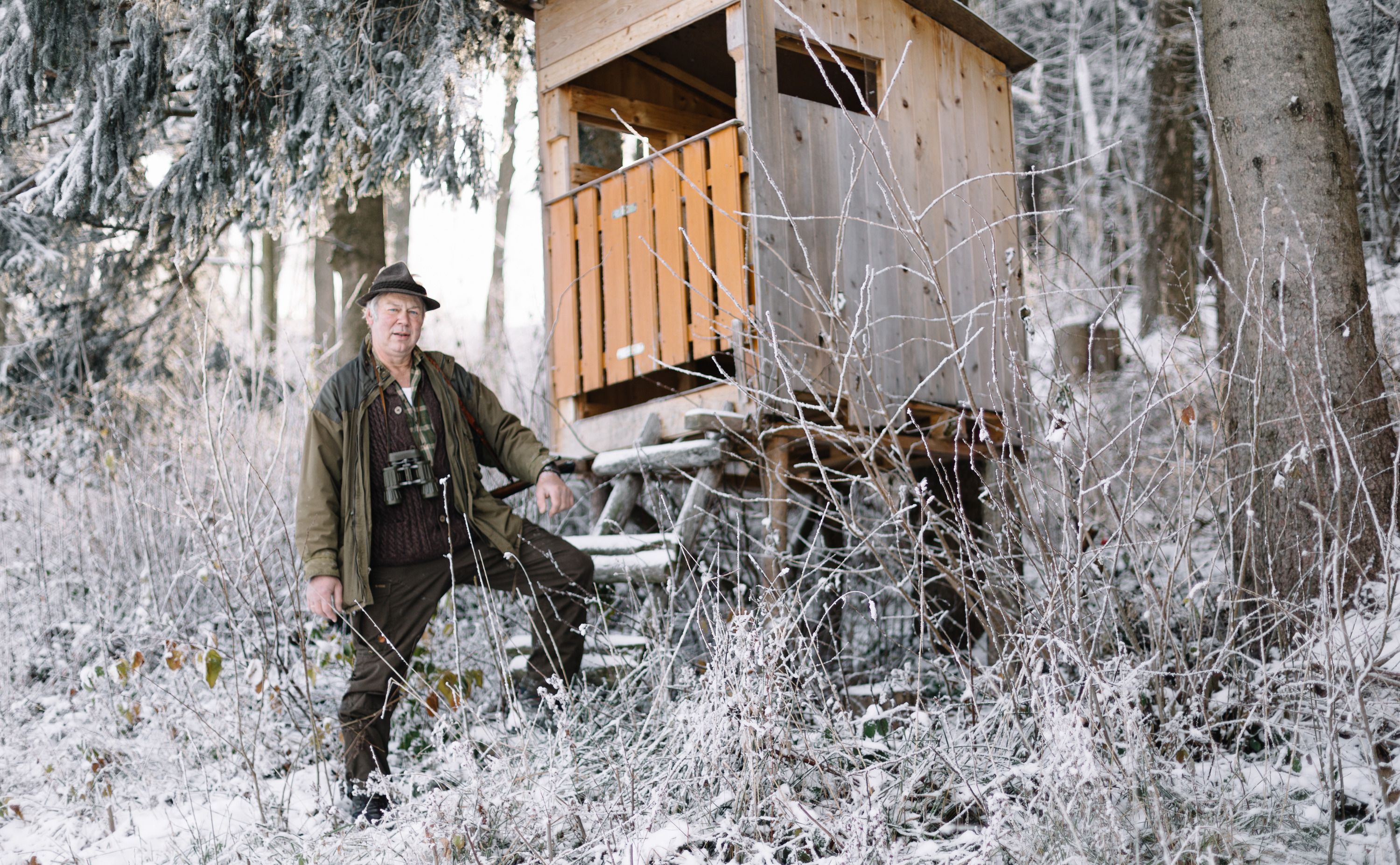 A man in hunting clothes stands next to a high seat in the snowy forest.