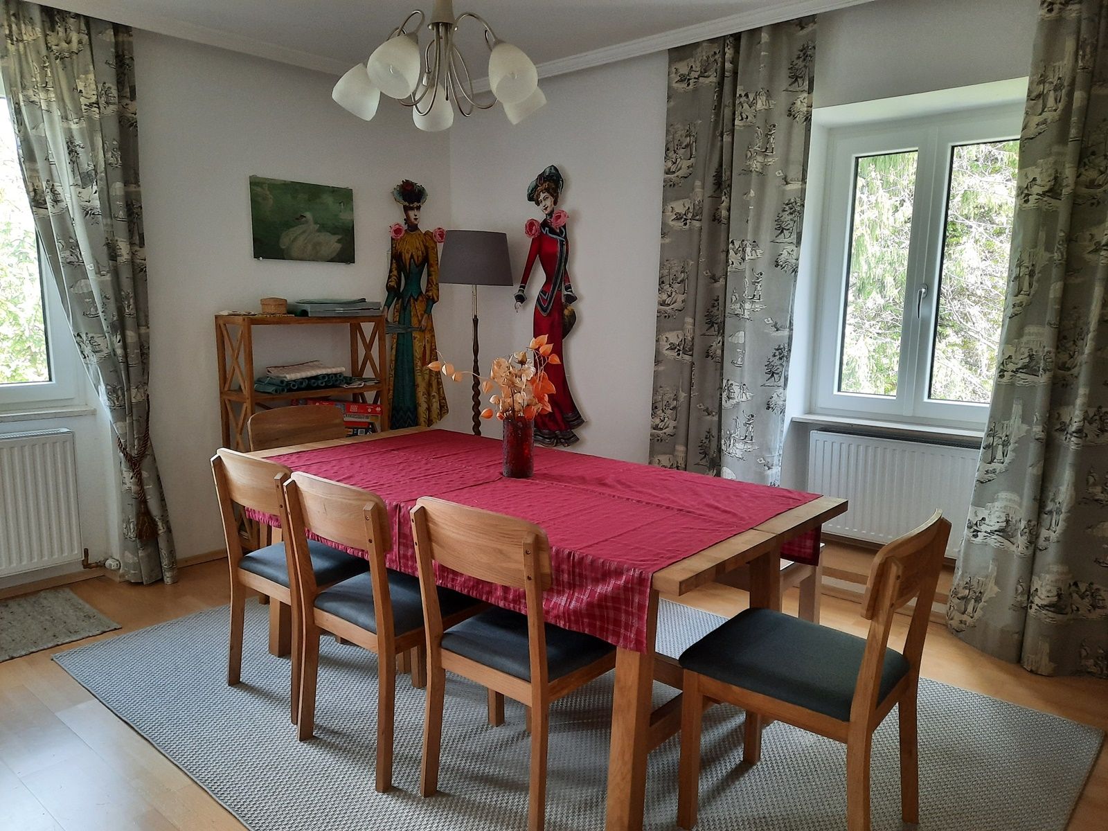 Dining area with wooden table, red table runner, wooden chairs, curtains and decorations on the wall.