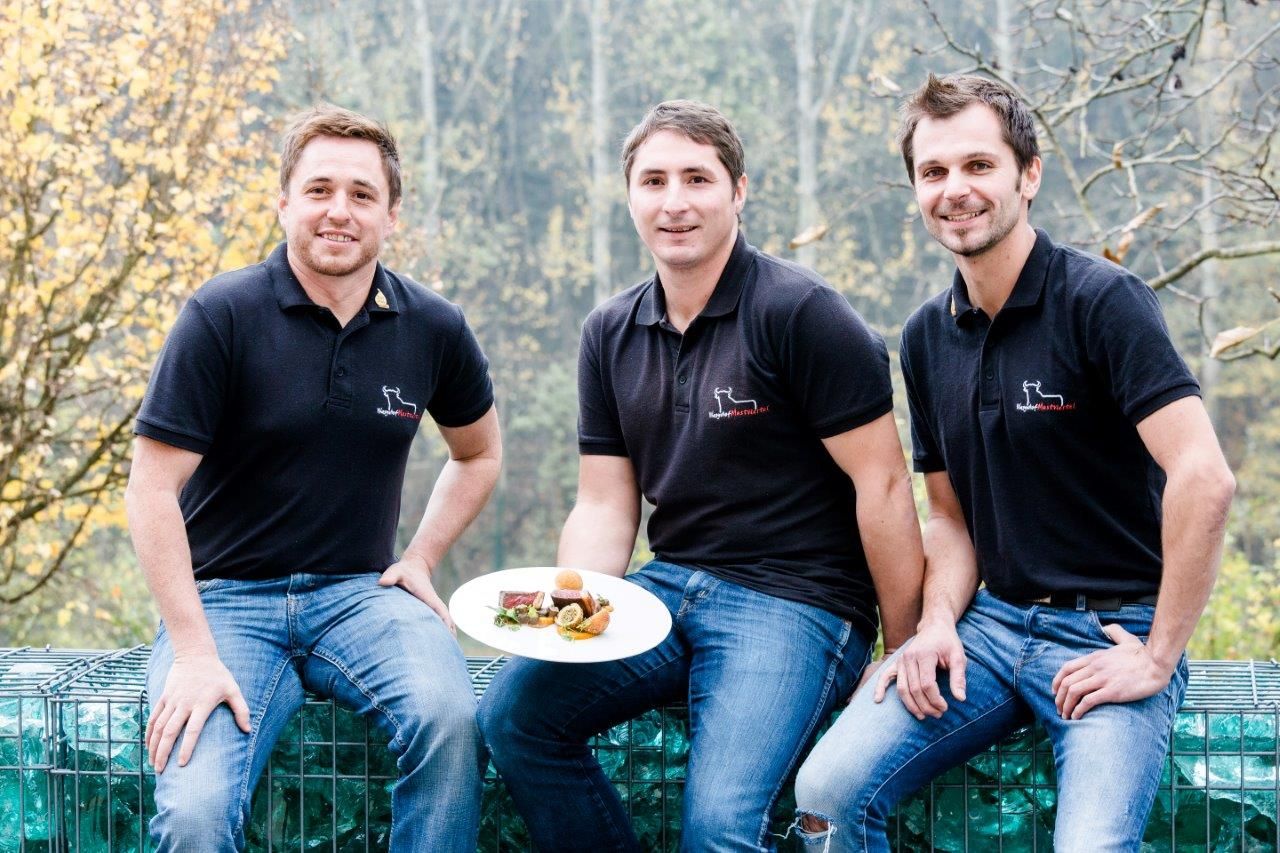 Three men in black polo shirts sit outside holding a plate of food.