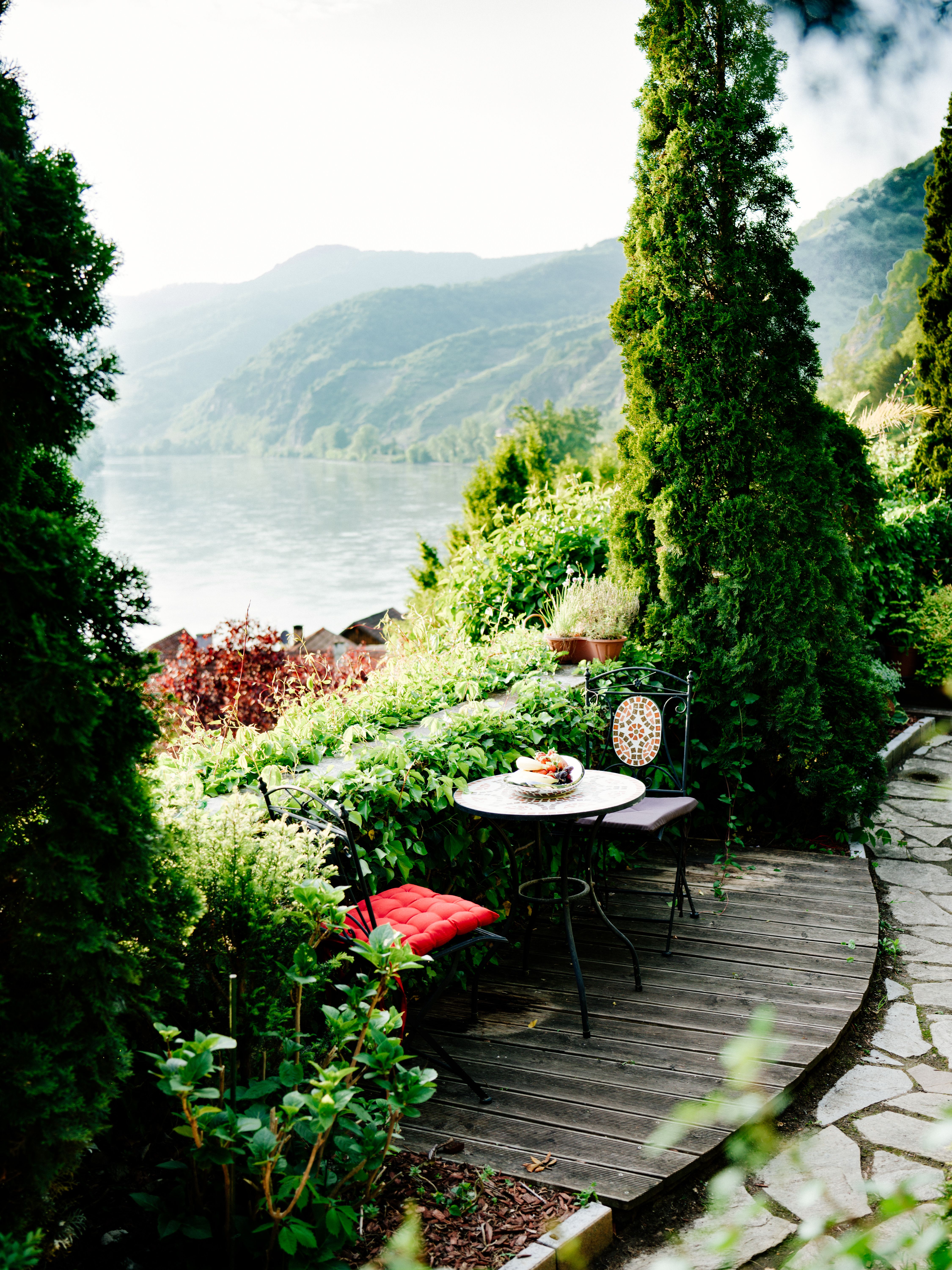 A small garden table with two chairs on a wooden terrace, surrounded by lush greenery and with a view of a river and wooded hills.