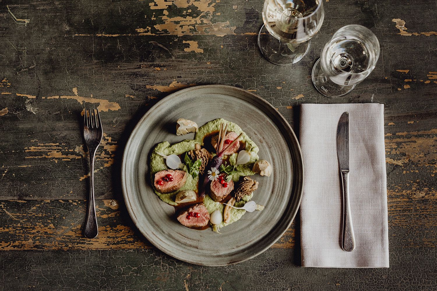 A plate of saddle of venison, coriander cream and vegetables on a rustic table.