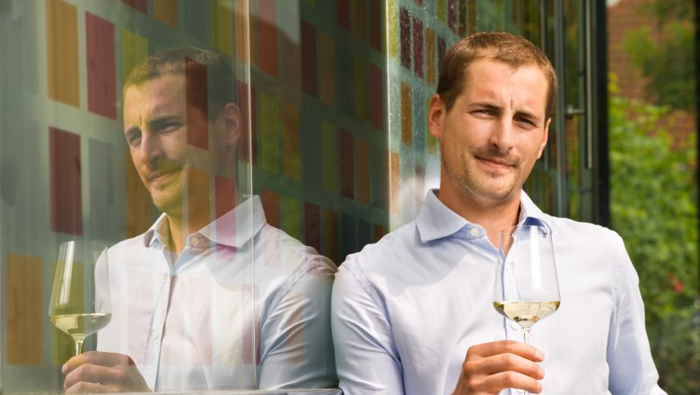 A man in a light blue shirt holds a glass of white wine and leans against a colorful glass wall.
