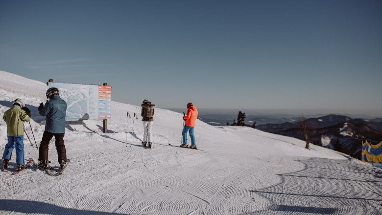 Skiers stand on a slope in the Unterberg ski area in front of a signpost, surrounded by snow-covered mountains and a clear sky.