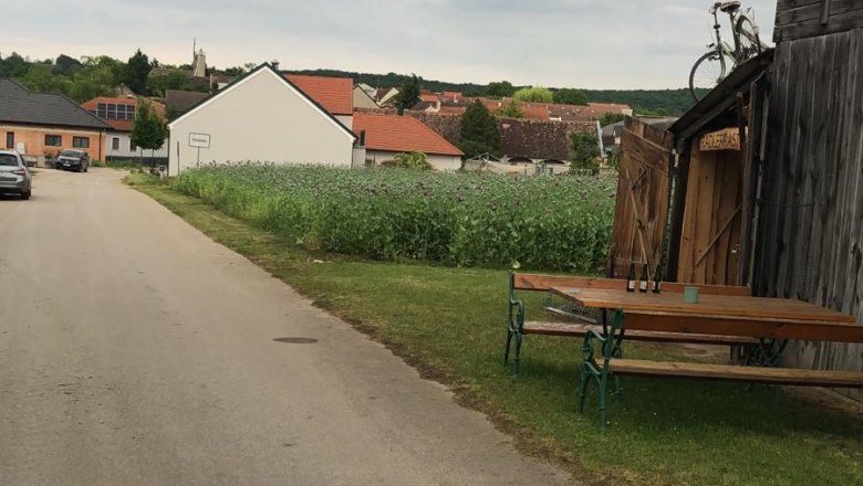 Village street with wooden hut and bicycle on the roof, benches and table next to it, fields and houses in the background.