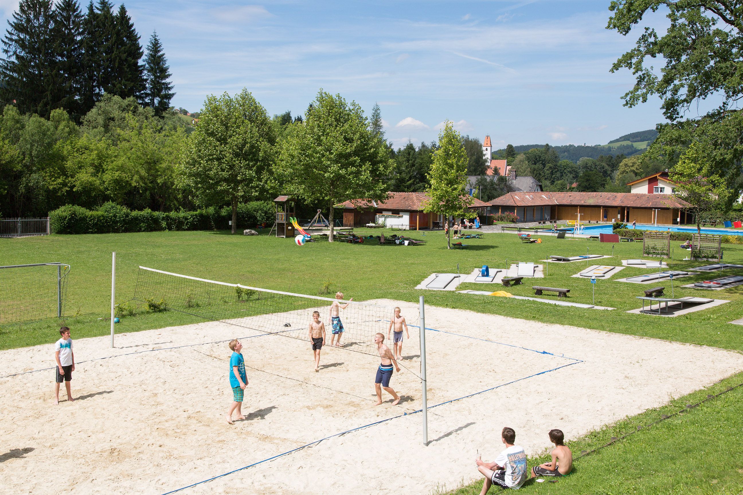 Children play volleyball on a sand court at the Aspang outdoor pool, surrounded by green meadows and trees.