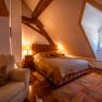 Cozy bedroom with wooden beams, large bed, armchair and carpet. Warm light from bedside lamps, window with view to the outside.