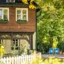 An idyllic country house with wooden elements, surrounded by flowering gardens and trees. There are two blue sun loungers in the garden.