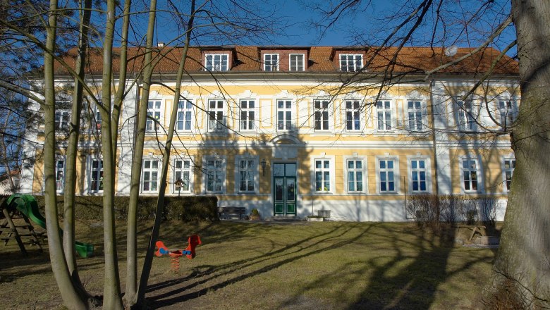 Yellow building with white window frames and red roof, surrounded by trees and a playground.