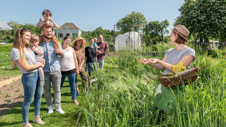 A group of people stand in a garden while a woman shows plants with a basket.