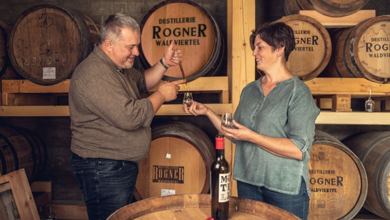 Two people at a tasting in a distillery with wooden barrels in the background.