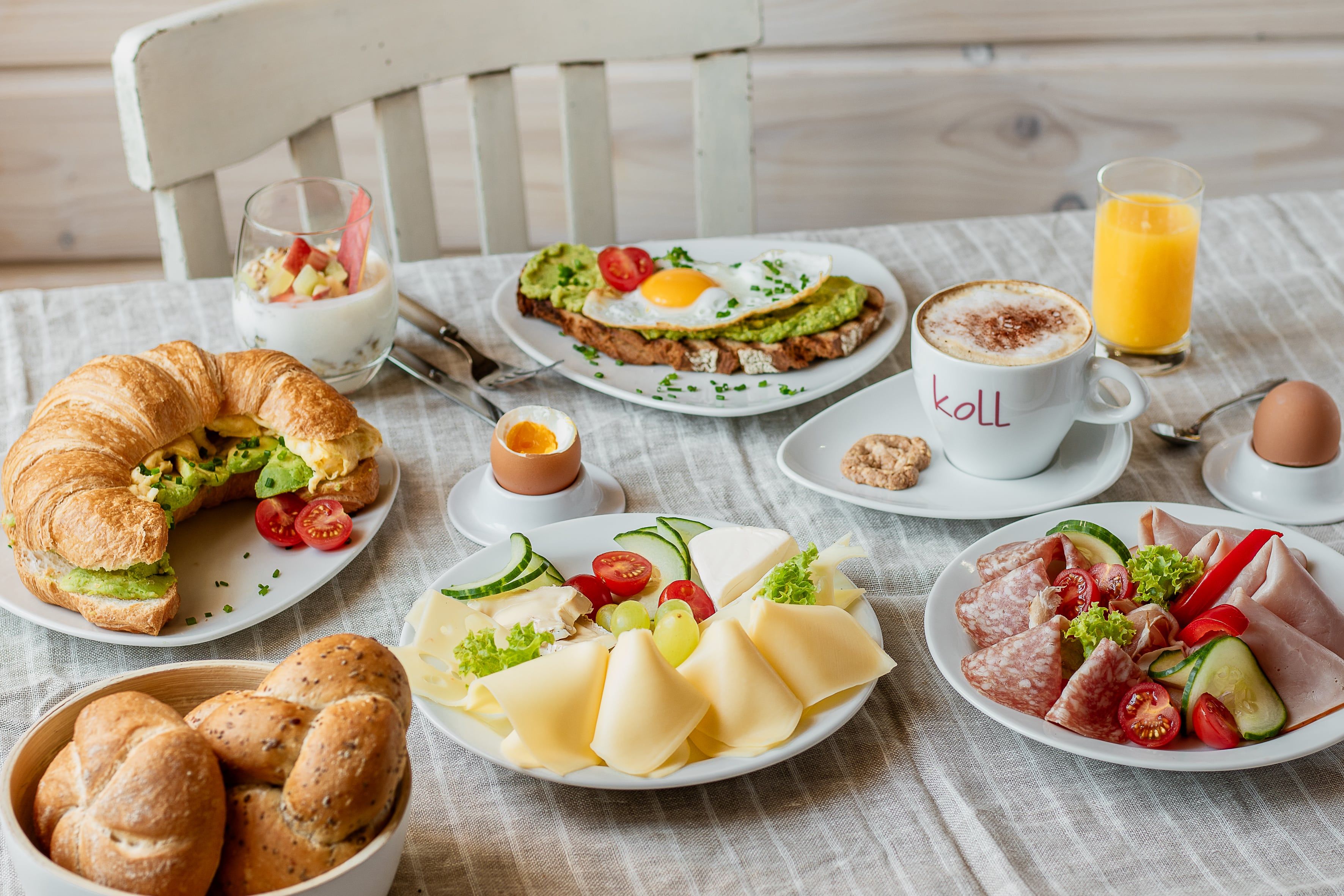 A hearty breakfast with croissant, bread rolls, cheese, cold cuts, avocado toast, yogurt, cappuccino, orange juice and soft-boiled egg.
