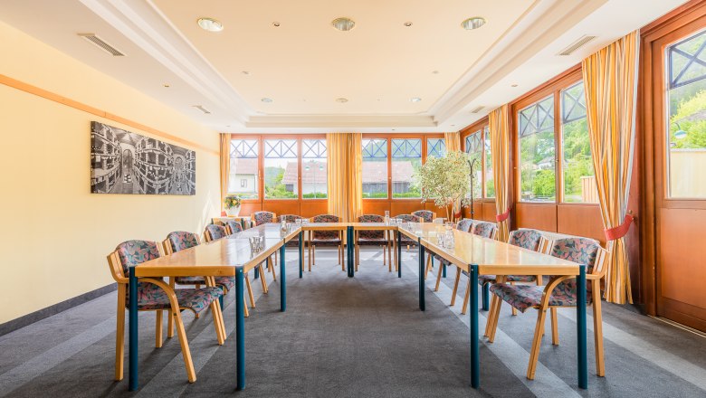 A bright seminar room with U-shaped tables, chairs and large windows with curtains.