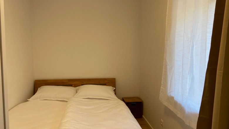 A simply furnished bedroom with a double bed, white sheets and a small bedside table. A window with a curtain is visible.