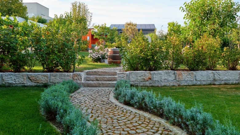 Well-kept garden with stone path, bushes and trees, in the background a building with solar panels.