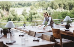 A waitress sets tables on a terrace with a view of green forests.