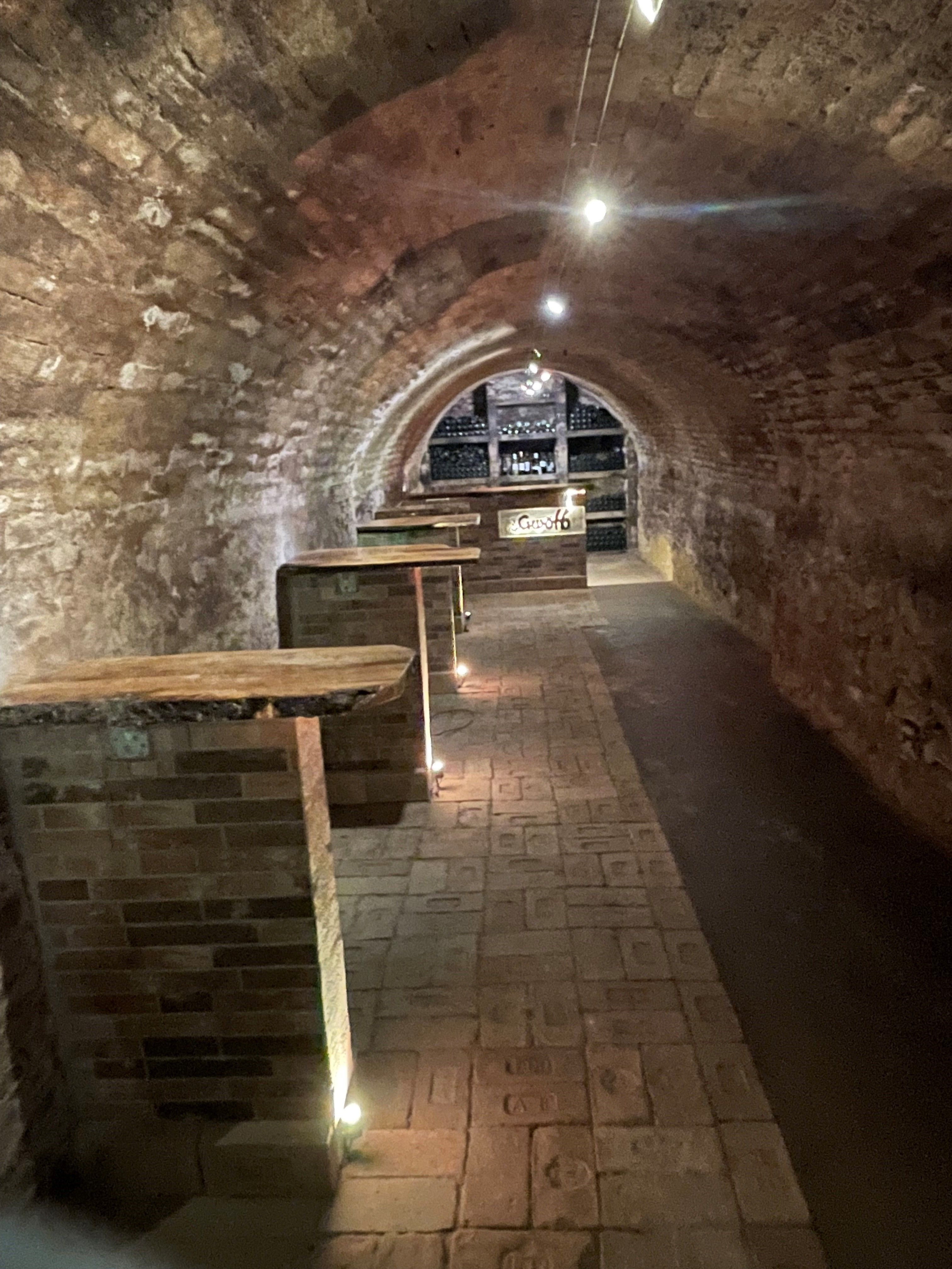A vaulted wine cellar with brick walls and floor, illuminated by small lights along the floor. Wine racks can be seen in the background.