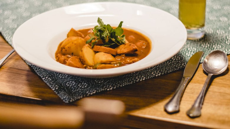 A plate of potato goulash on a table, cutlery and a glass of drink next to it.