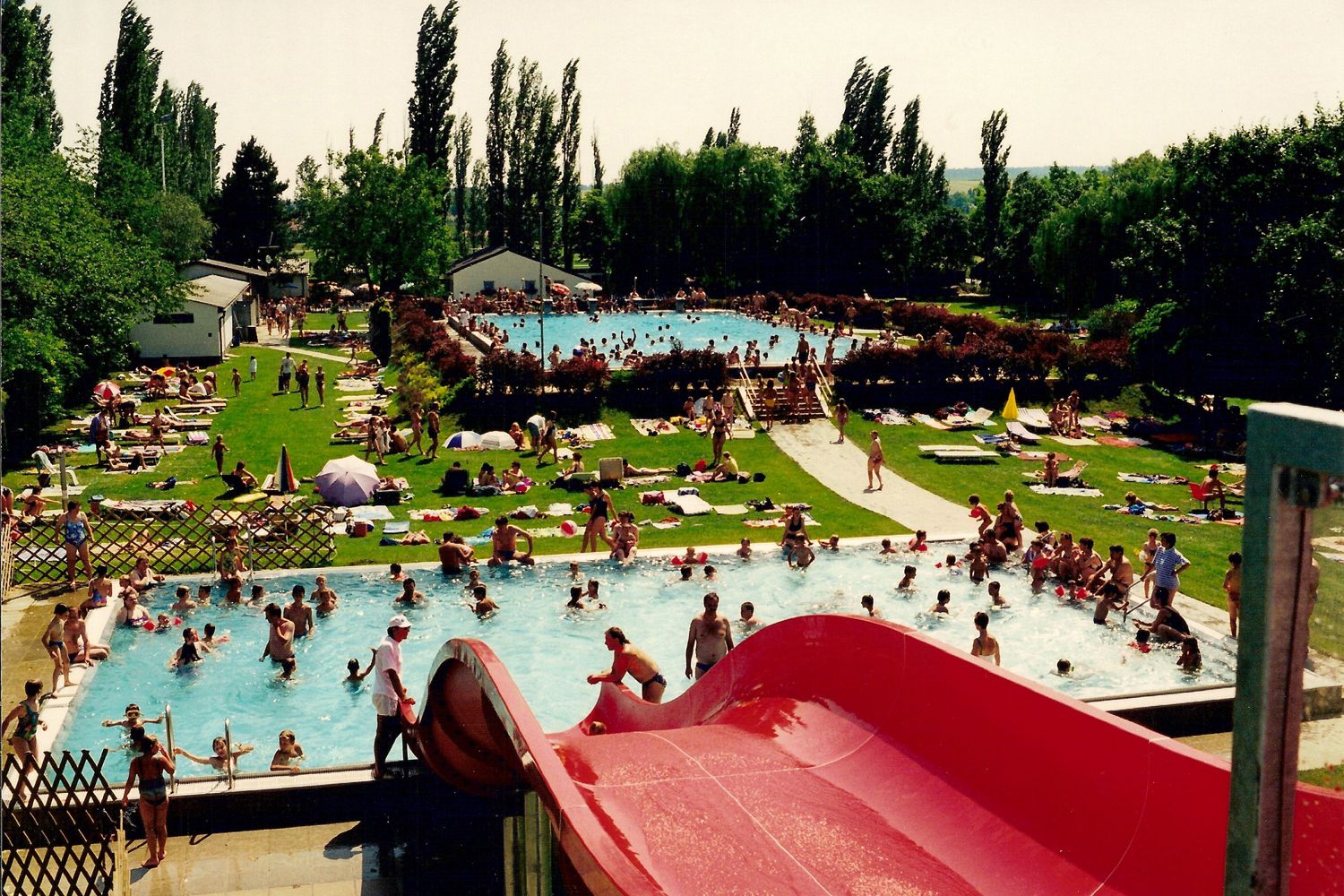 Outdoor pool with lots of people in the water and on the sunbathing lawn, red slide in the foreground.