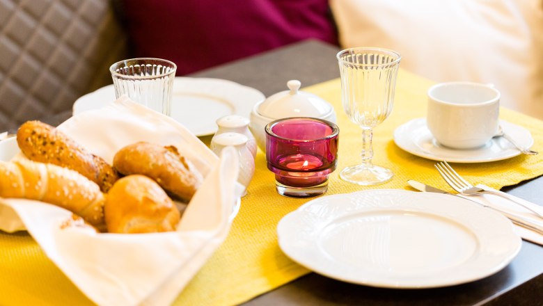 Breakfast table with bread basket, crockery and glasses on a yellow tablecloth.