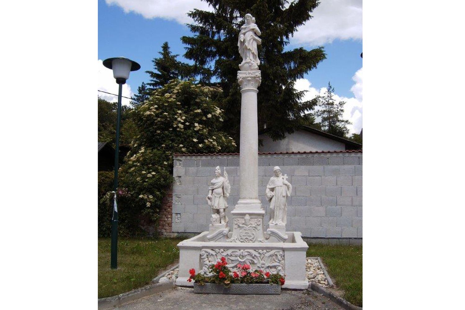 Marian column with statues and flowers, surrounded by trees and a wall.