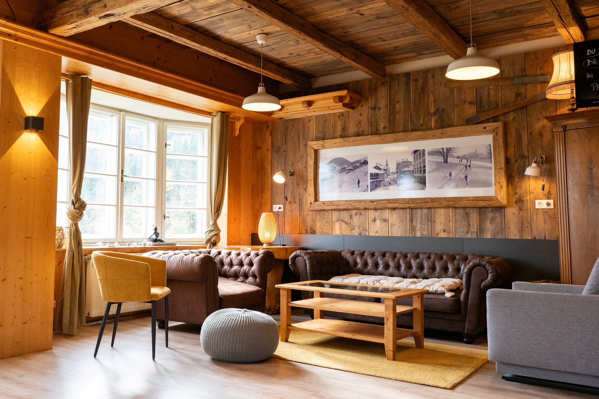 A cozy common room with wooden walls, leather sofas, and a large window.
