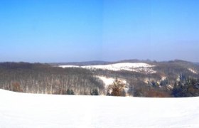 Snow-covered landscape with hills and forests under a blue sky.