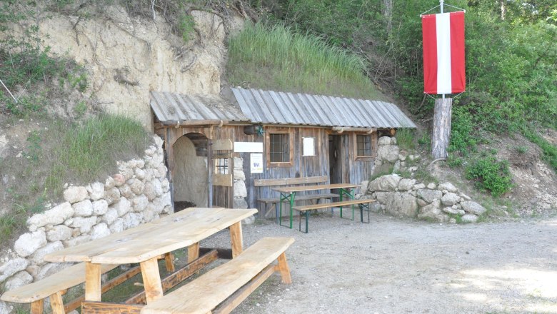 A rustic wooden hut with a table and benches in front of it, built into a hill. A red and white flag hangs from a tree.