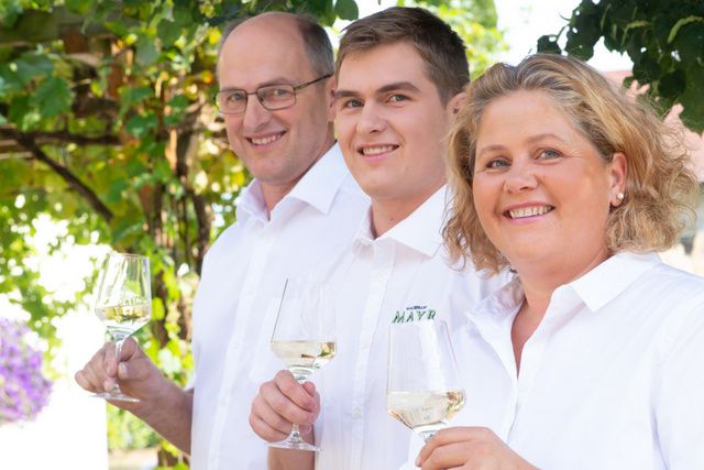 Three people in white clothing hold glasses of wine and smile at the camera.