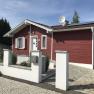 Red wooden house with white shutters and solar panels on the roof, surrounded by a paved courtyard and a low wall.
