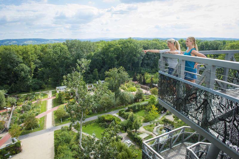 Two women stand on a lookout tower and look out over a green garden with trees and paths.