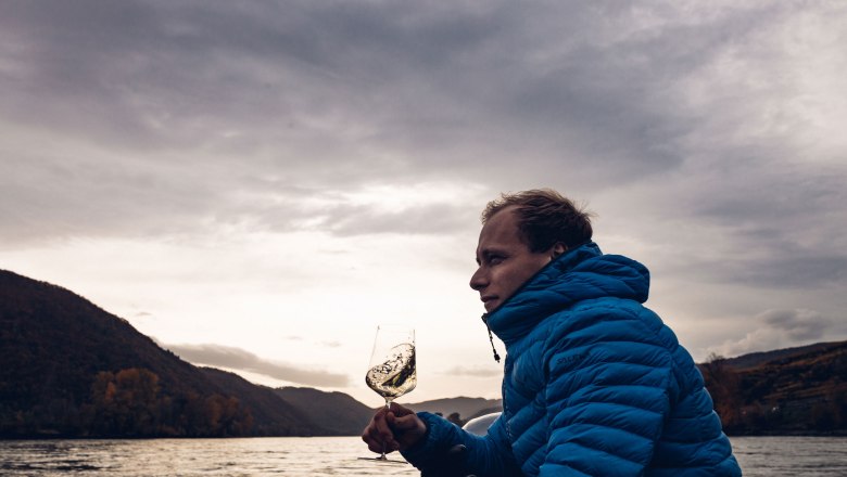 Person in blue jacket holding a wine glass on a boat in front of a river landscape.