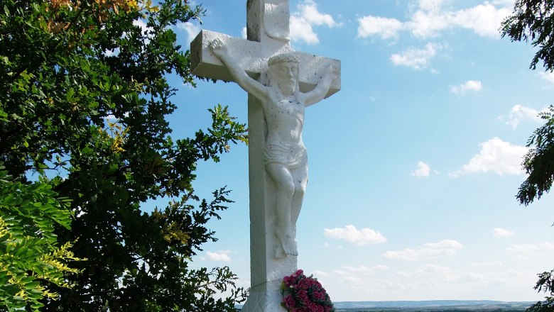 White stone cross with figure of Jesus and wreath of flowers, surrounded by trees and landscape in the background.