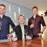 Three men are standing at a table with wine glasses and bottles of wine.