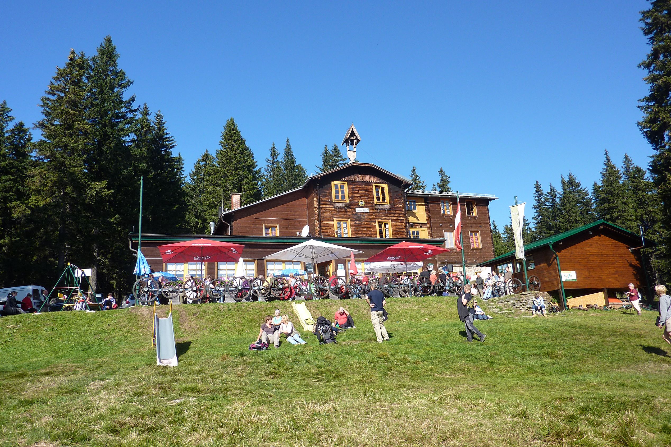 Wooden house in the mountains with people and bicycles in front of it, surrounded by trees.
