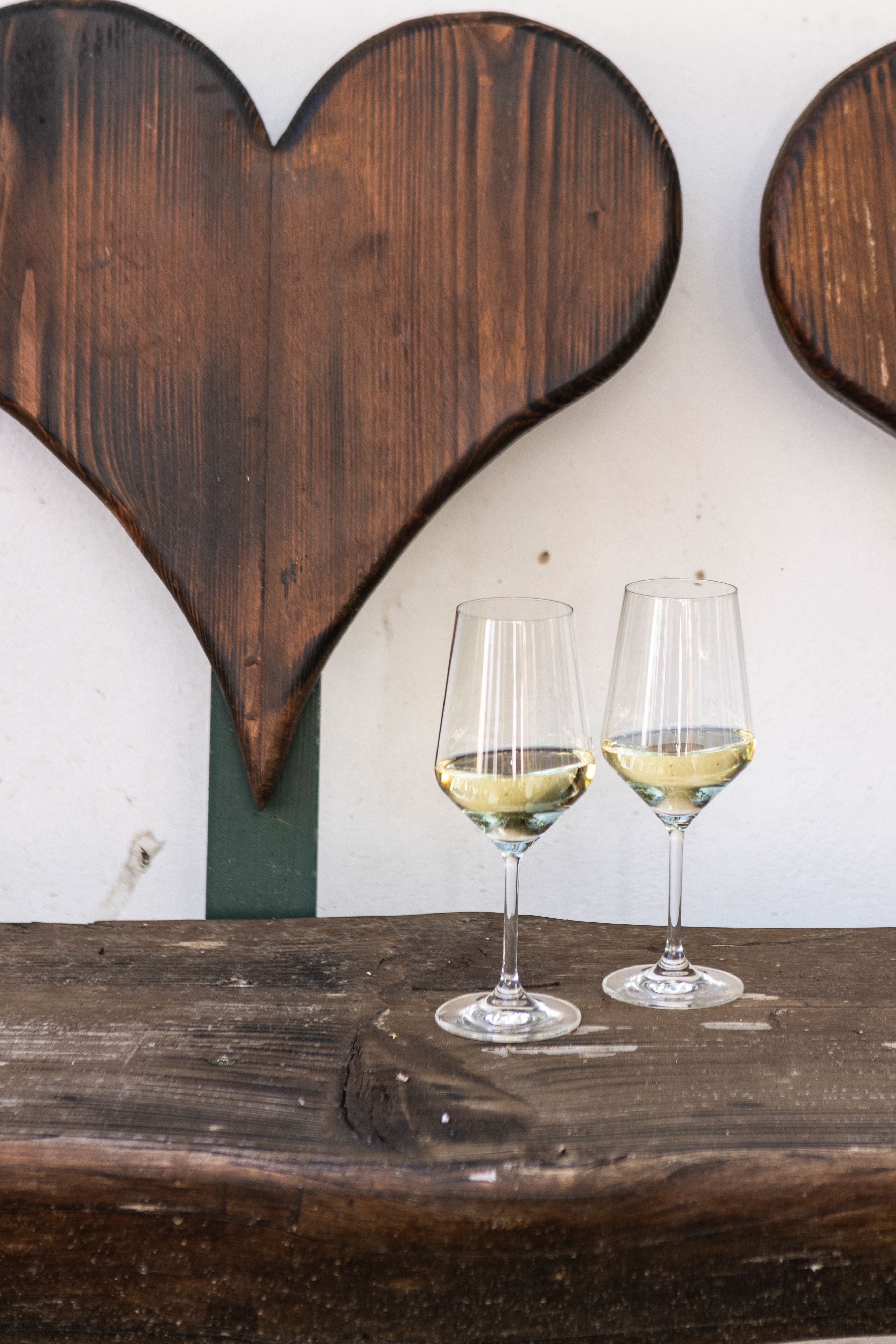 Two wine glasses with white wine on a wooden table in front of a wooden wall with heart decoration.