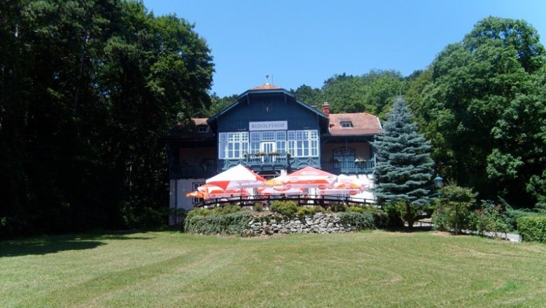 A large house called Rudolfshof with a terrace and sunshades, surrounded by trees and a meadow.