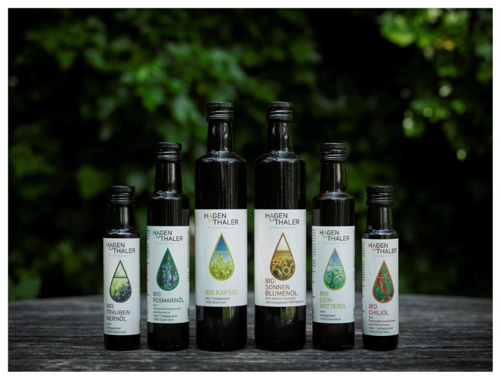 Six bottles of Hagenthaler organic oils on a wooden table against a green background.