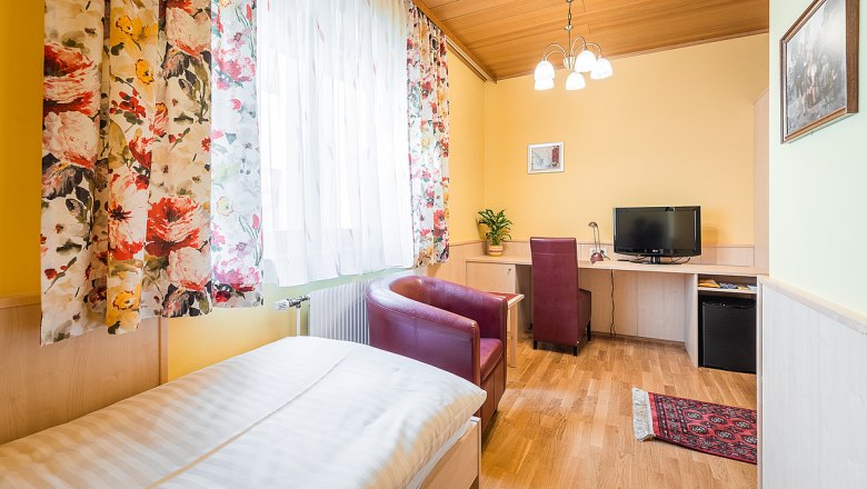 A cozy single room with a wooden floor, a bed, a desk with chair, a TV and colorful curtains.