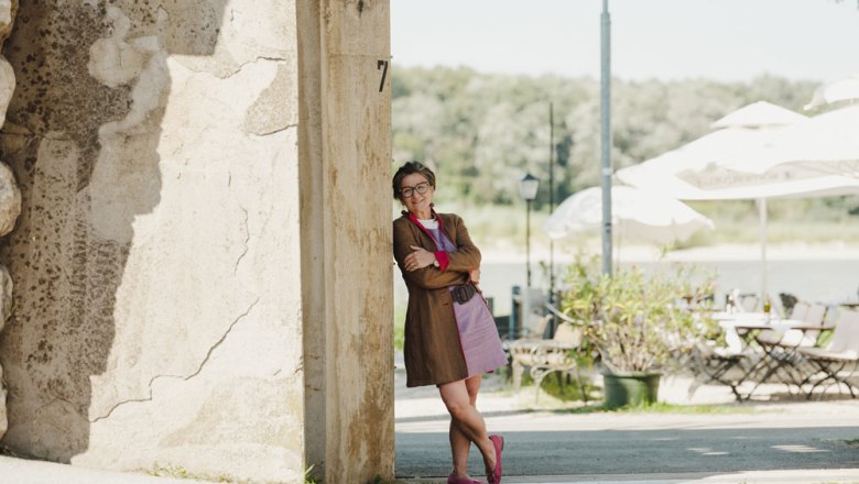 Landlady Michaela Brenner-Melchior, © Niederösterreich Werbung/Michael Reidinger Smiling woman leaning against a wall, in the background a terrace with tables, chairs and parasols.