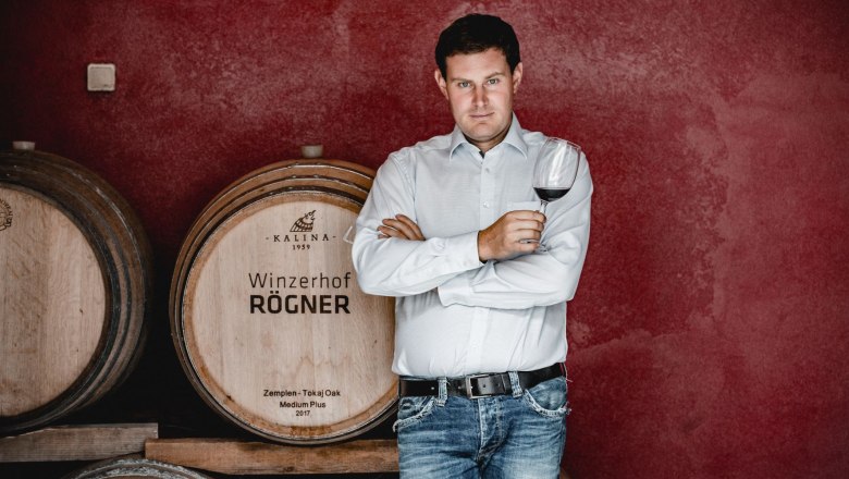 A man stands in front of wine barrels and holds a glass of red wine in his hand.