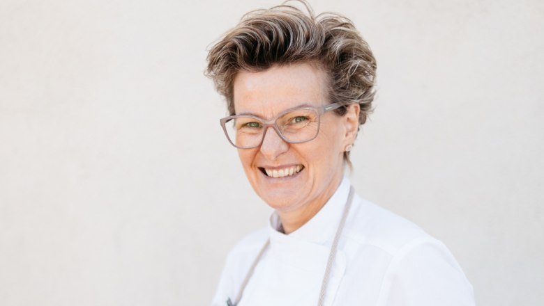 A smiling woman in chef's clothes holds a basket of fresh vegetables.