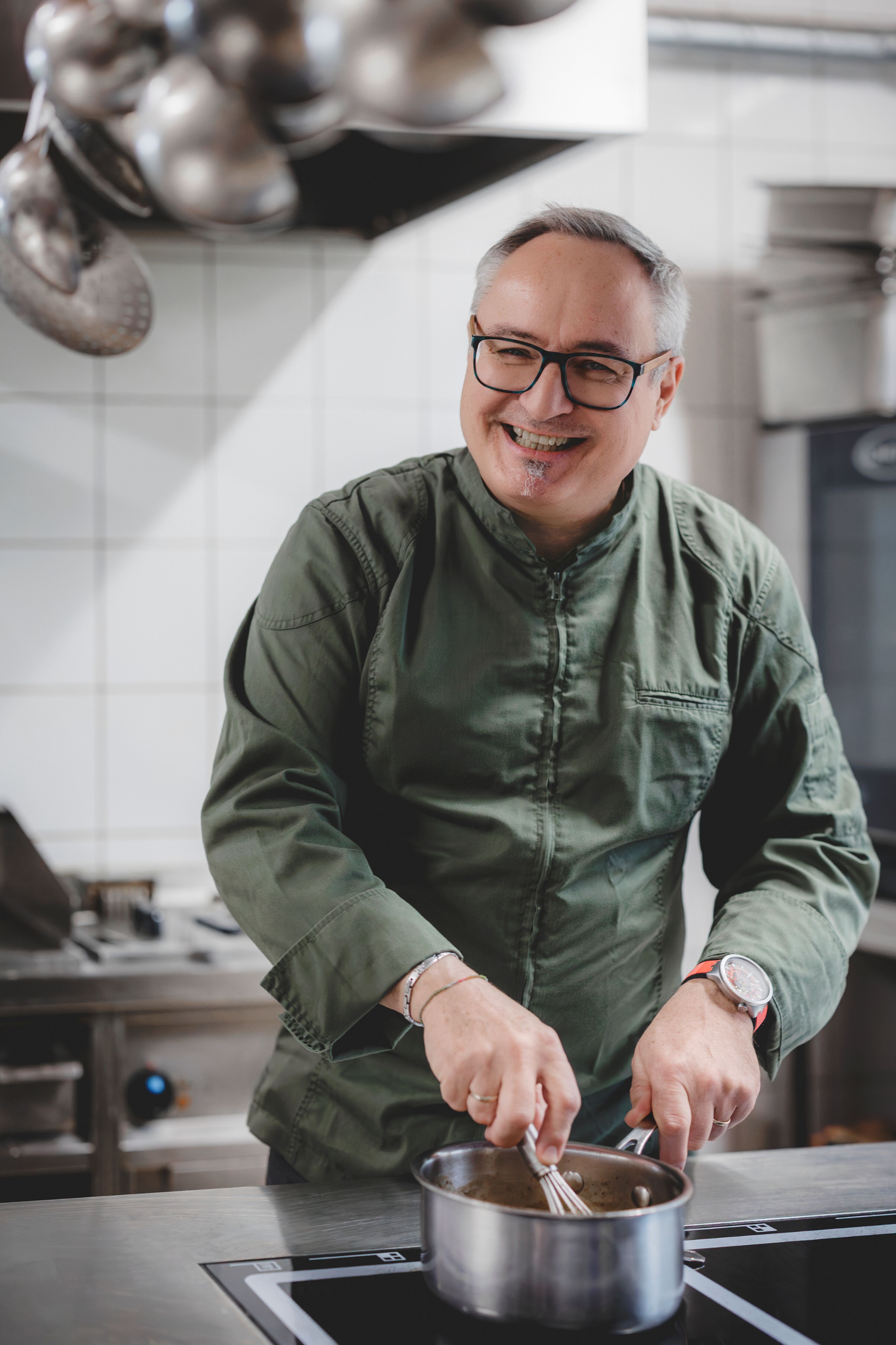 A smiling chef in a green jacket stirs a pot in a professional kitchen.