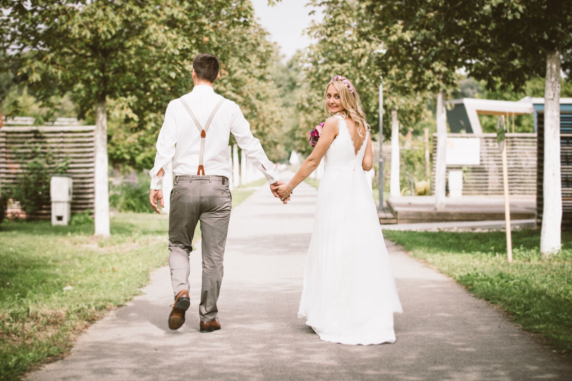 A bride and groom walk hand in hand along a path in the garden, the bride smiles back.